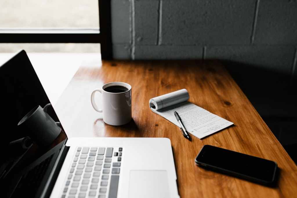 A table with a laptop, notepad, cup of coffee, and phone